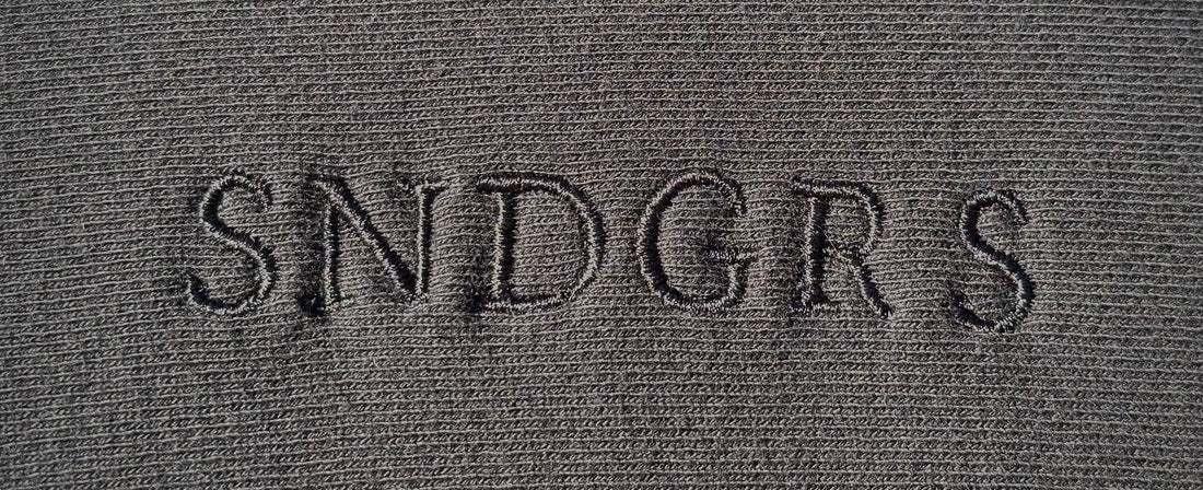 Close-up of tonal SNDGRS center-chest embroidery on black heavyweight fleece.