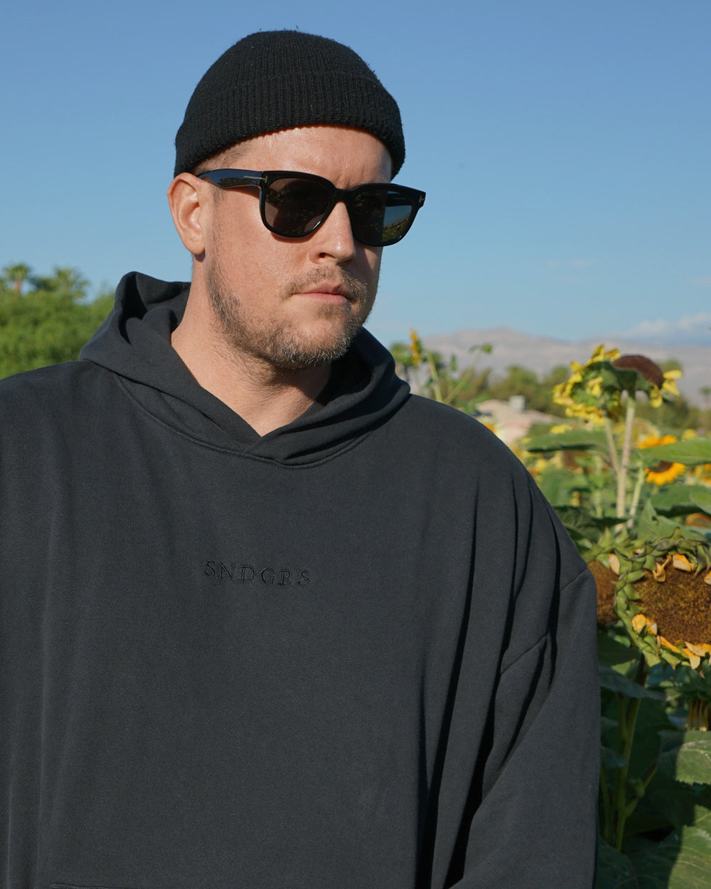 An adult wearing sunglasses and a black beanie stands outdoors in a black SNDGRS hoodie, with tonal embroidery across the chest and sunflowers behind.