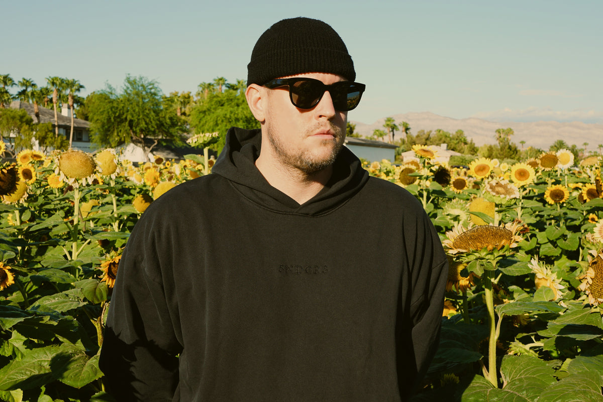 SNDGRS founder wearing the black STILL. Volume 001 hoodie with tonal chest embroidery in a sunflower field.